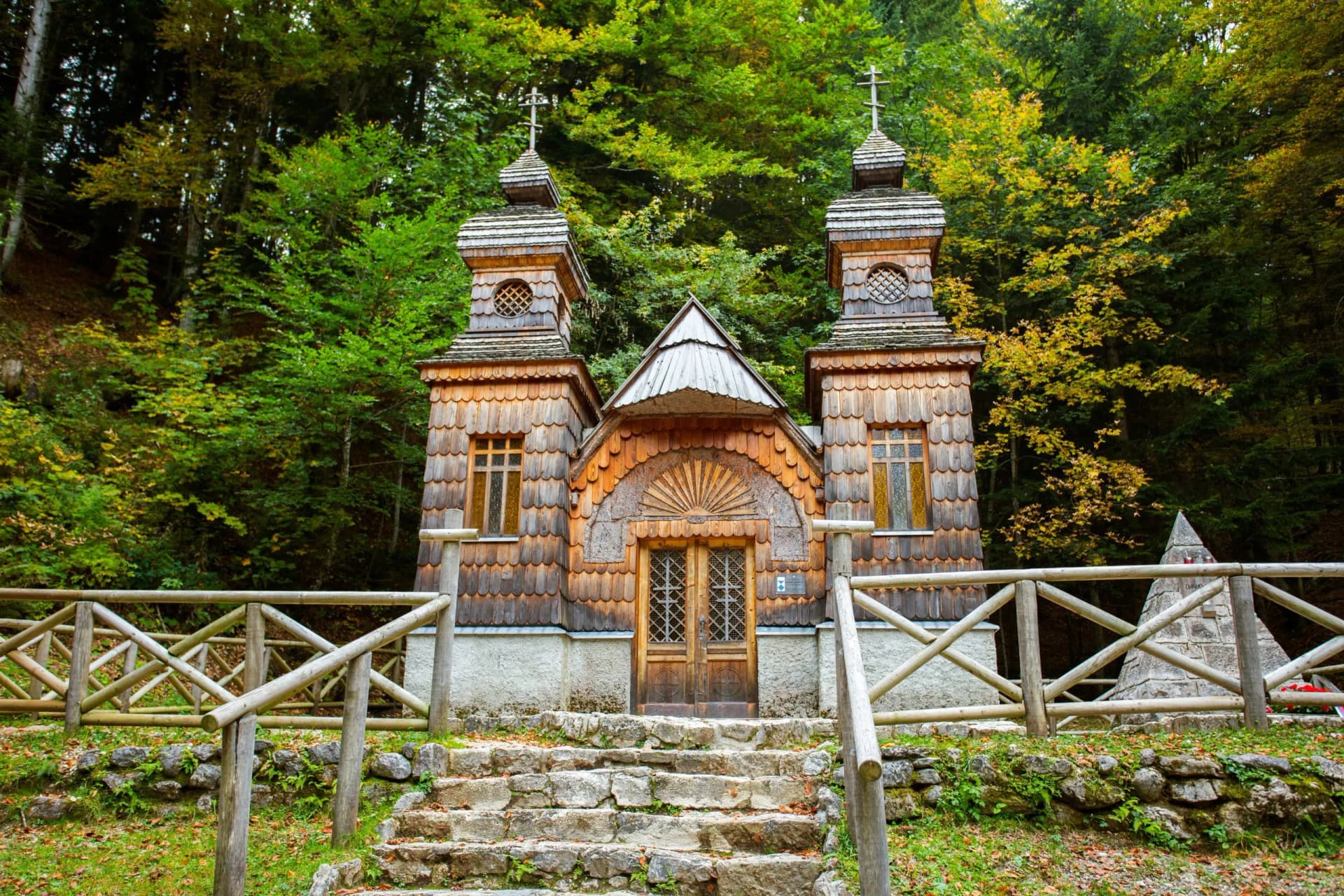 Wooden chapel with shingle siding and twin towers set against a dense forest with early autumn colors.