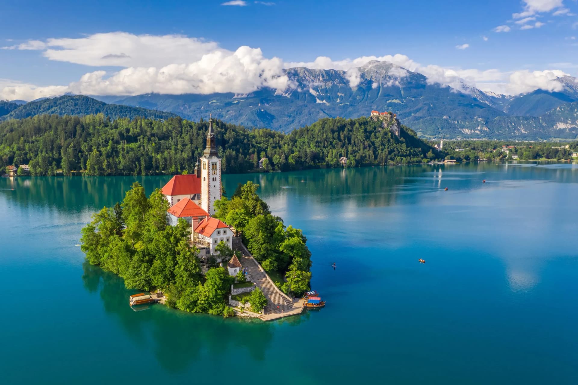 Lake Bled island church surrounded by green trees on turquoise water with mountains in background.