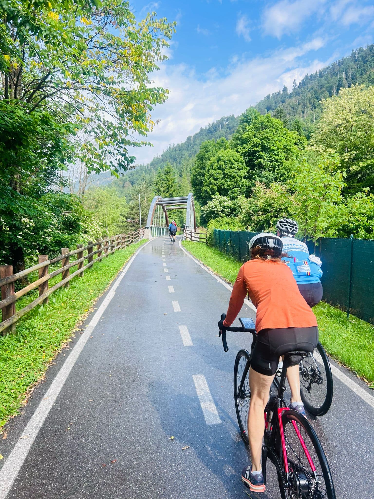Cyclists riding on a wet paved path toward a small bridge in Kranjska Gora with forested mountains.
