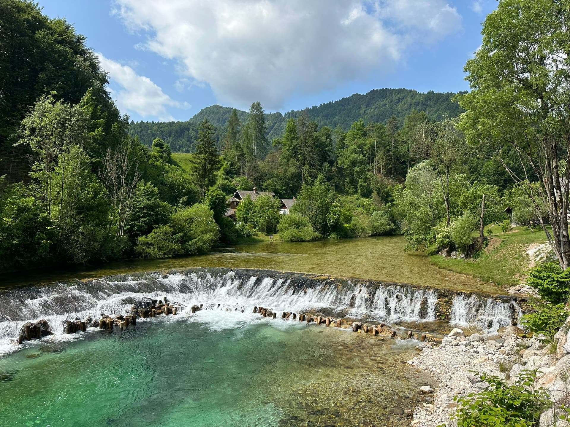 Waterfall over low weir on turquoise river with forested mountains and houses in background