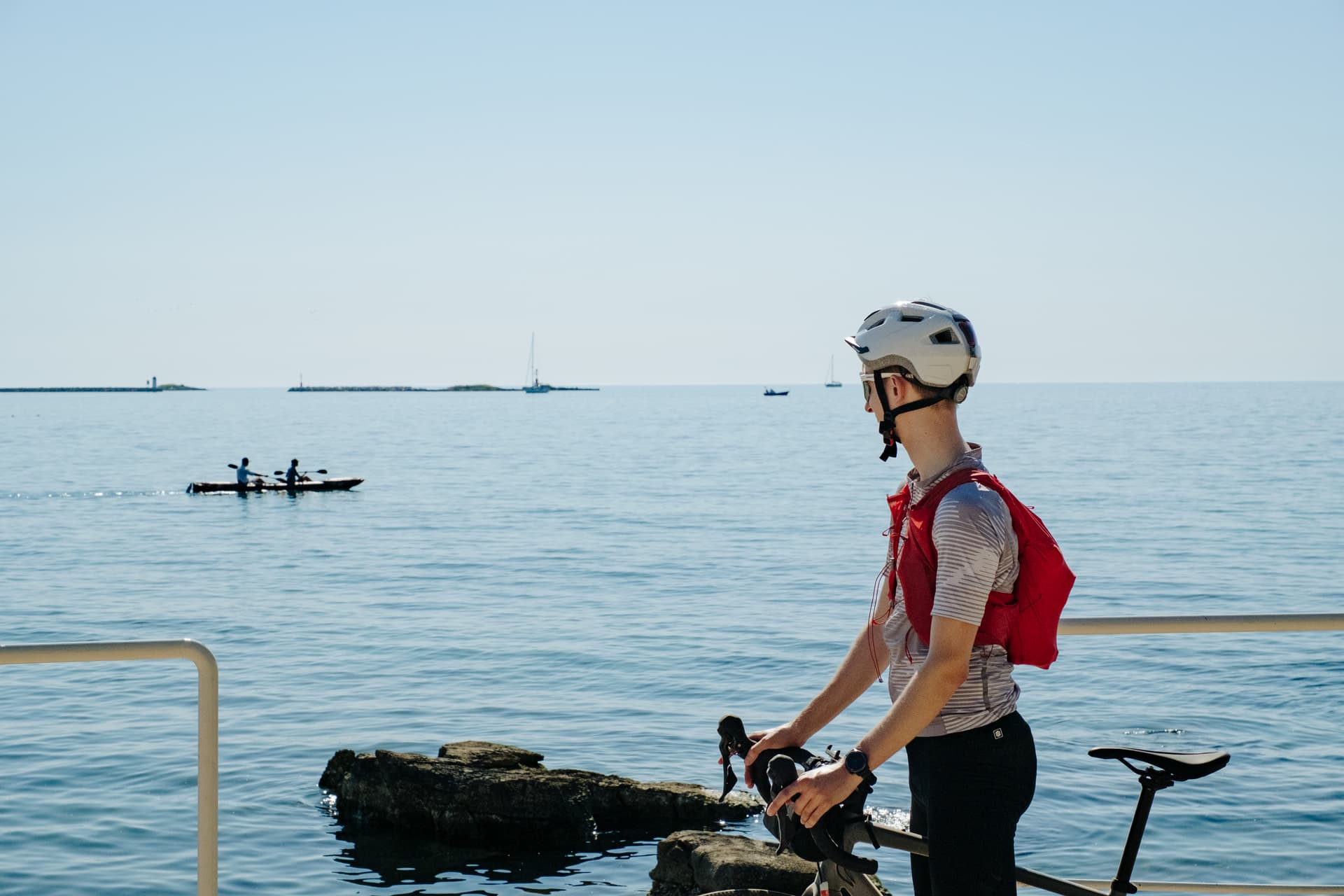 Cyclist with helmet and red pack pauses by the water with kayaks and sailboats visible.