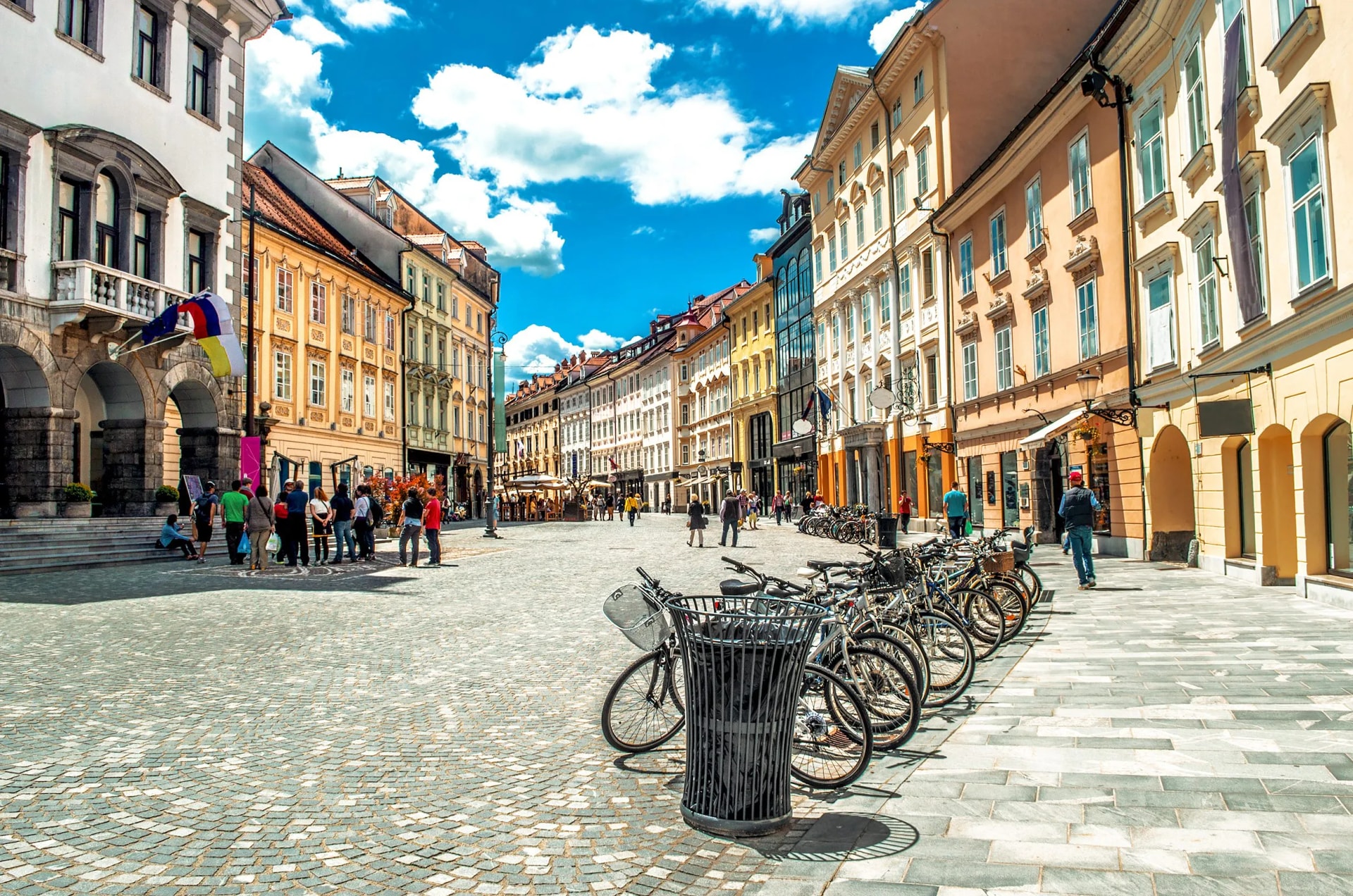 Cobblestone square with historic buildings, parked bicycles, and people under a bright blue sky.