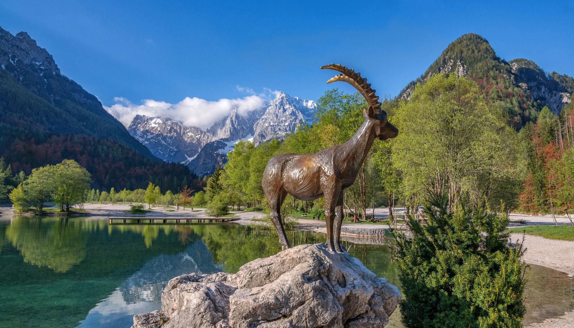 Bronze Zlatorog statue on rock by alpine lake with snow-capped mountains.