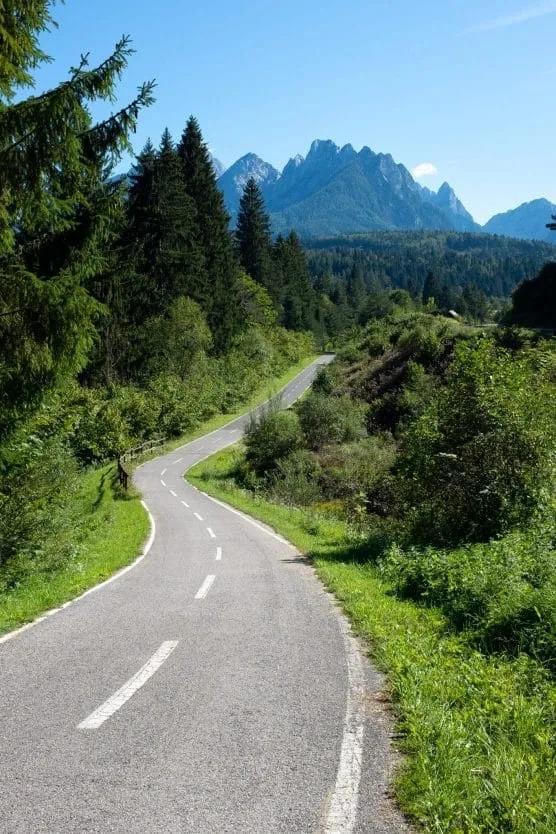 Winding paved road through lush green forest with jagged mountain peaks in background