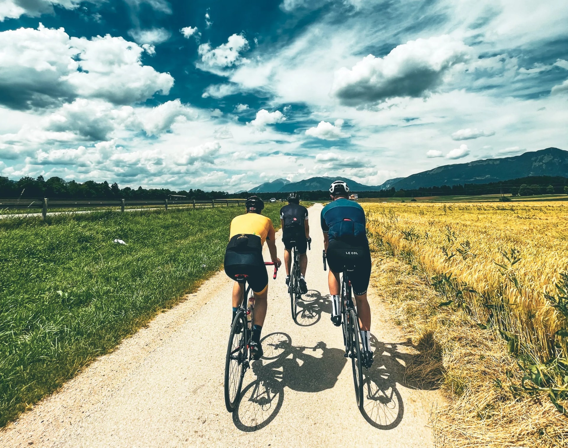 Three cyclists ride on a dirt road between green grass and golden fields toward mountains under a dramatic cloudy sky.