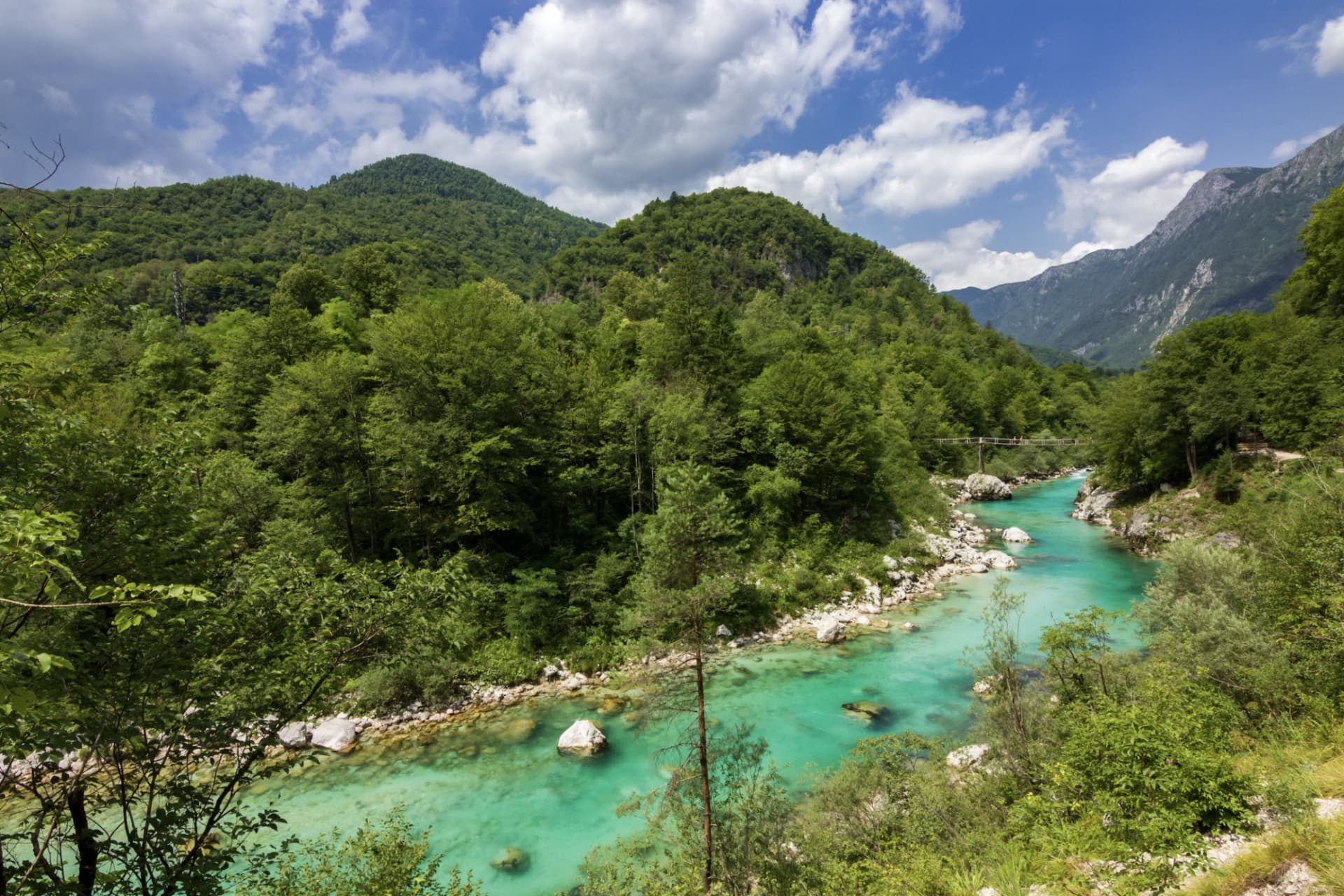 Turquoise river flowing through lush green valley with forested mountains under a blue, cloudy sky.