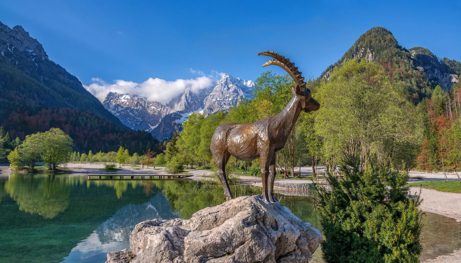 Bronze Zlatorog statue on rock by alpine lake with snow-capped mountains in background.