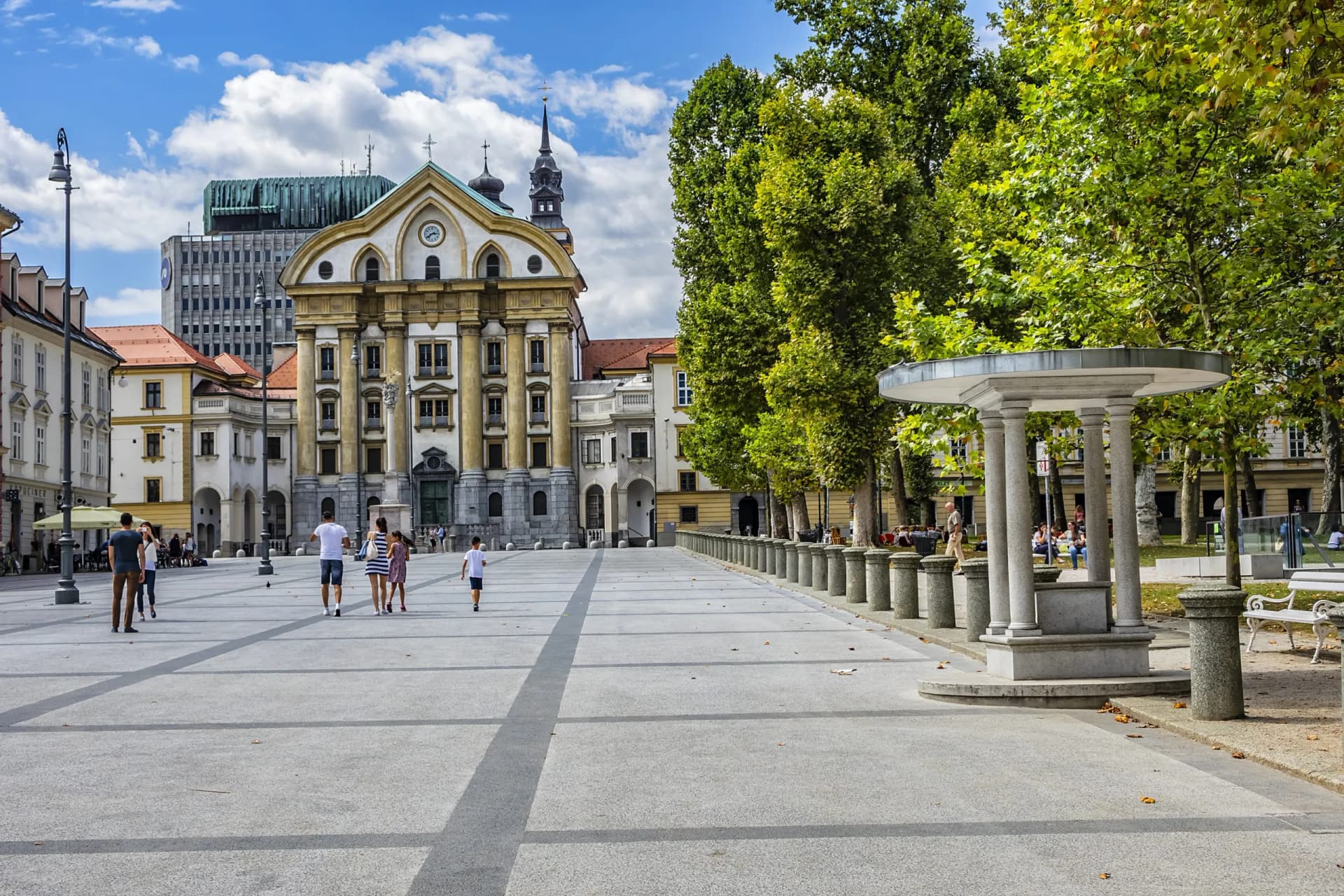 Kongresni Square in Ljubljana with historic architecture, trees, and people walking on a sunny day.
