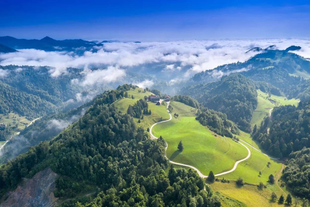 Rolling green hills above clouds near Polhov Gradec, with a winding road leading to a hilltop farm.