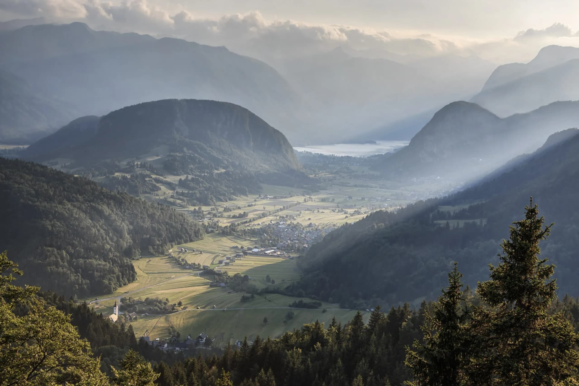 Misty alpine valley with village, lake, and layered mountains at sunrise or sunset.