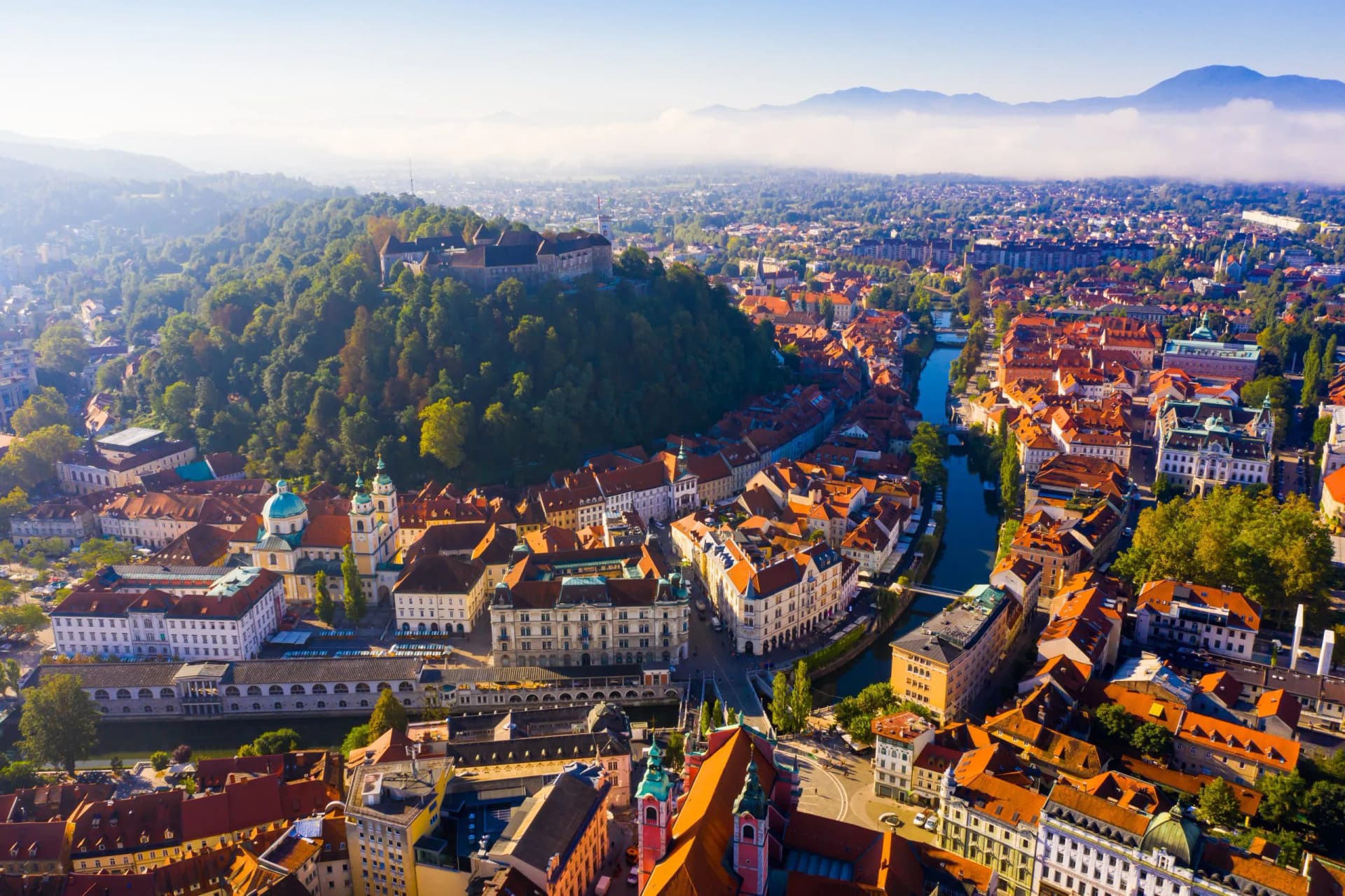 Aerial view of Ljubljana city with castle on forested hill and mountains in background.
