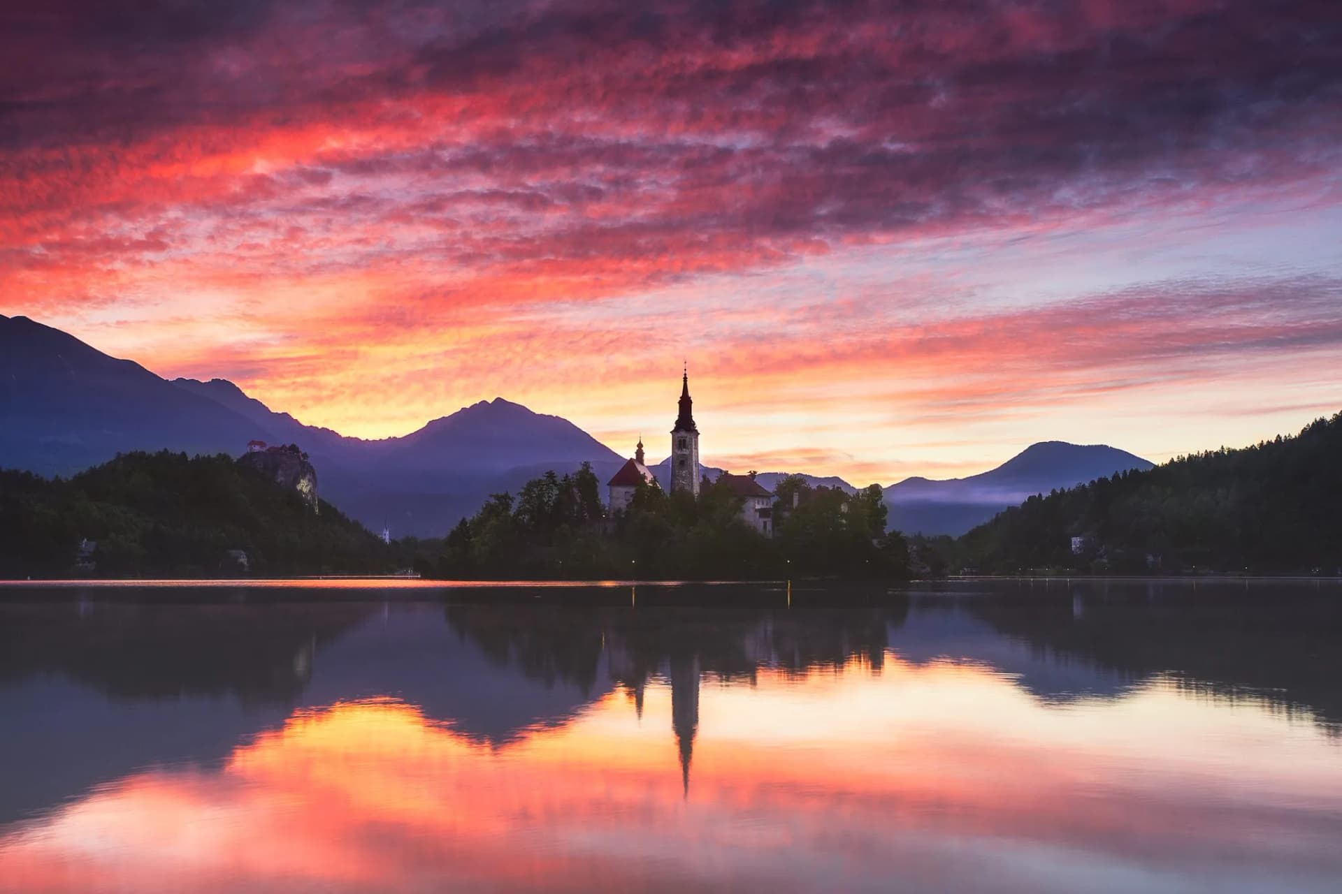 Lake Bled island church reflected in water at vibrant sunrise with mountain silhouettes.
