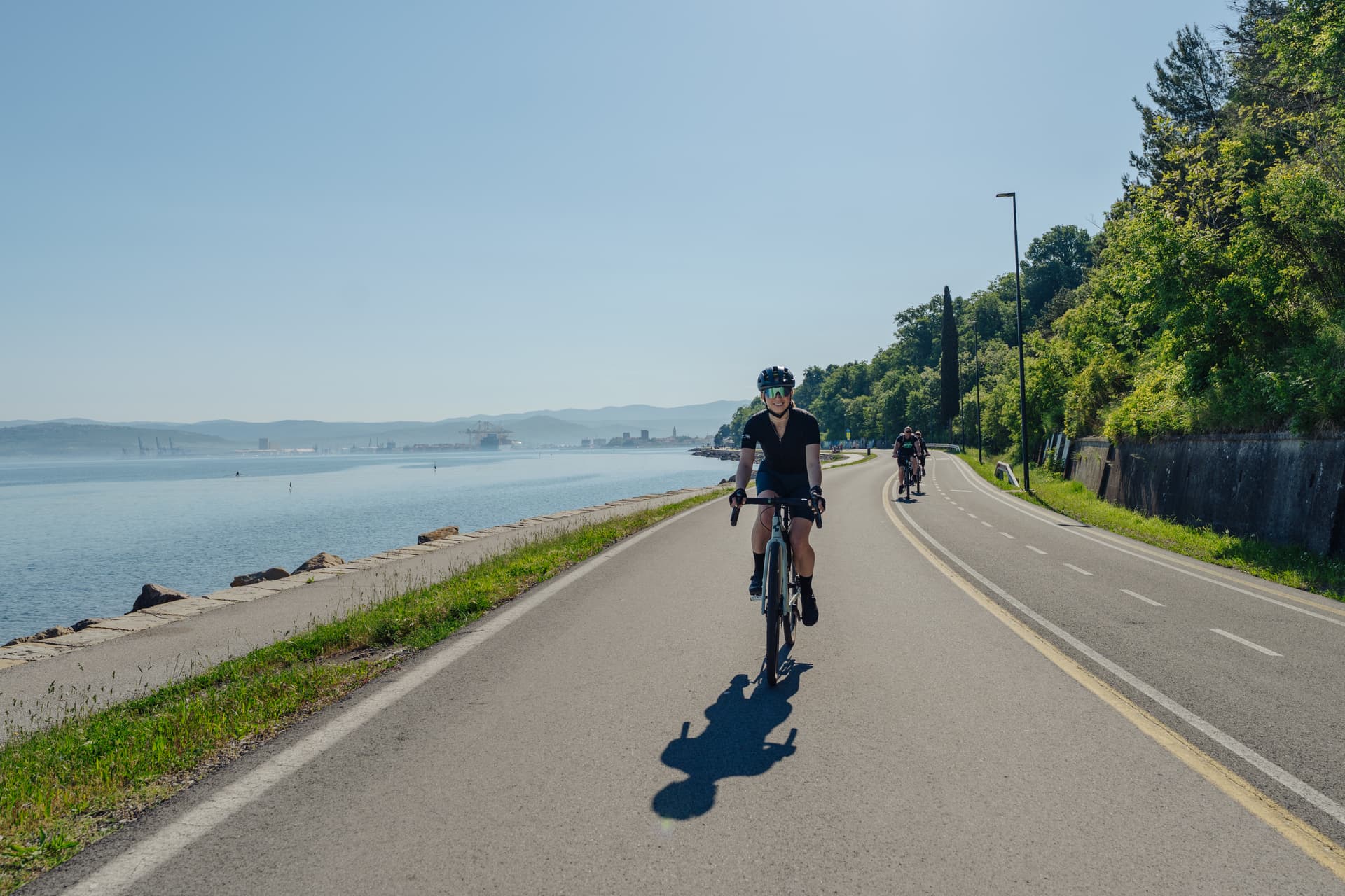 Cyclists riding on paved coastal road next to the sea with distant port cranes and hills.
